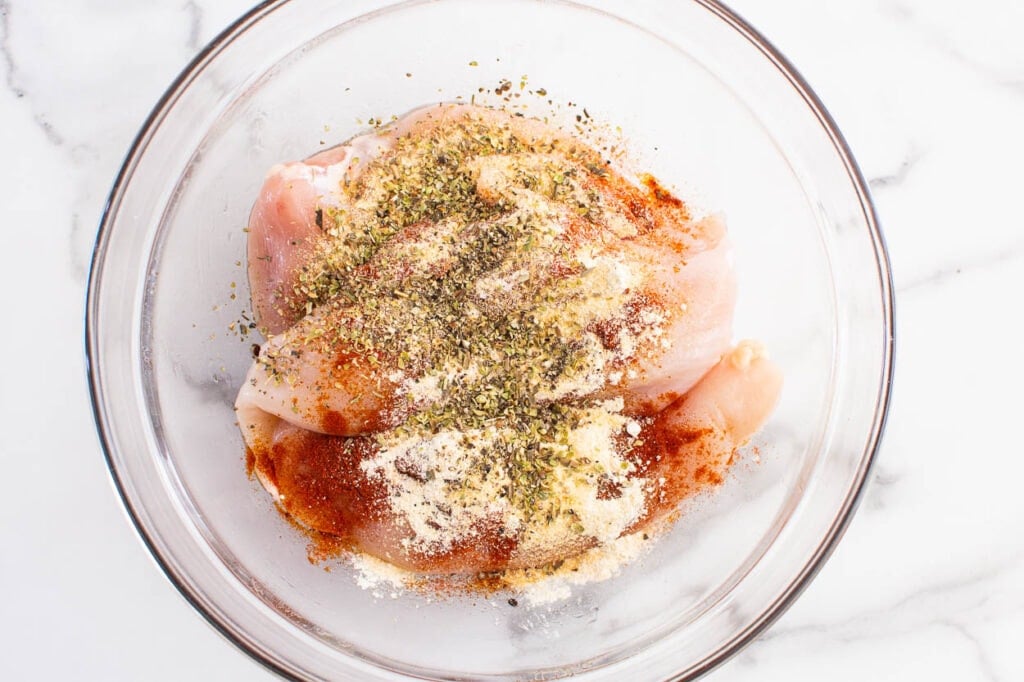 Chicken breasts coated with seasonings in a glass bowl.