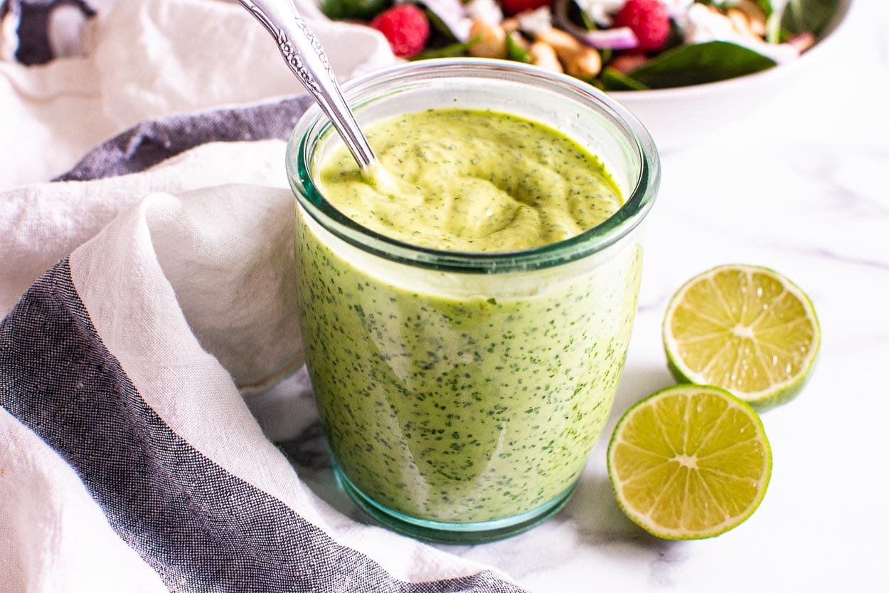 Avocado cilantro dressing in jar with a spoon and limes, a linen and salad.