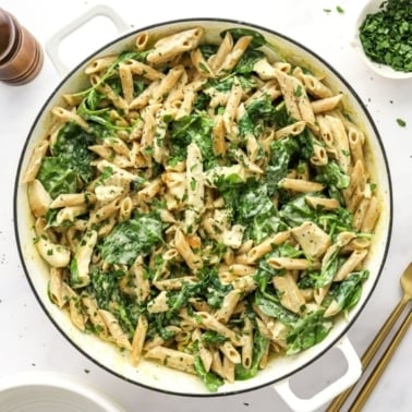 Chicken spinach pasta in a white pot, pepper grinder and small bowl of chopped parsley.