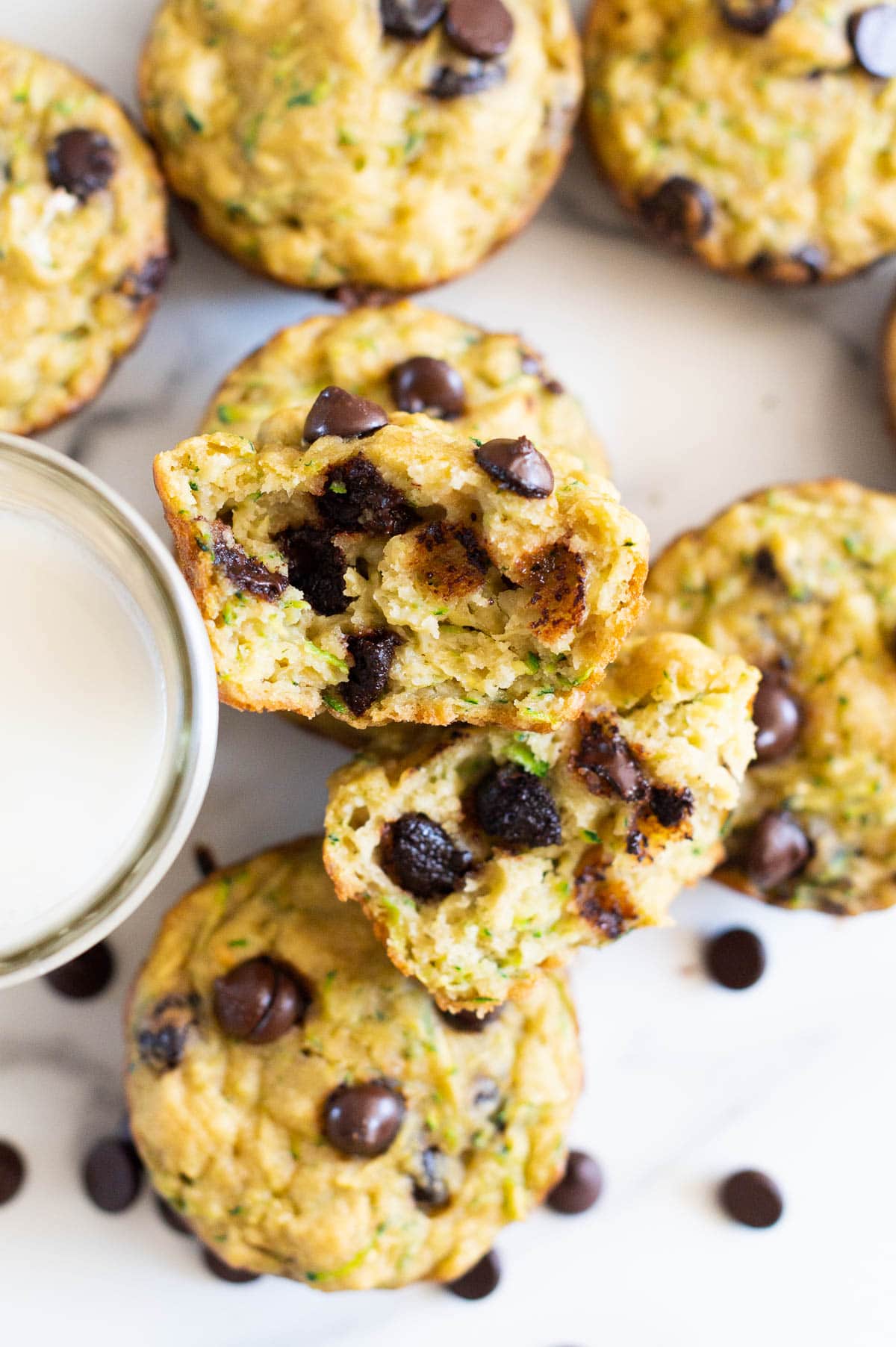 Chocolate chip muffins on a countertop at one muffin showing texture inside. A glass of milk.