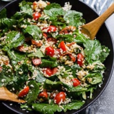 Baby kale salad with rice and grape tomatoes in black bowl with wooden spoons.