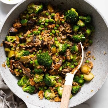 Broccoli mushroom stir fry with peas and brown rice in a skillet with a spoon.
