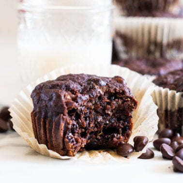 Chocolate zucchini muffin showing texture inside and more muffins and chocolate chips around it.