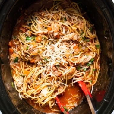 Crockpot chicken spaghetti with cheese and parsley on top and tongs inside.