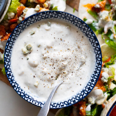 Greek yogurt blue cheese dressing in a bowl with a spoon. Buffalo chicken tacos around it on a board.