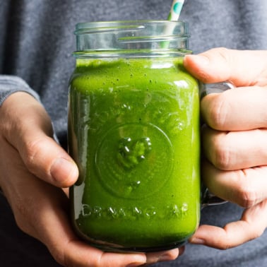 A person holding a jar with green smoothie and a straw.
