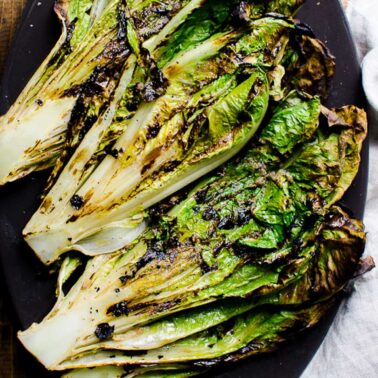 Grilled romaine lettuce served on black platter on wooden table. Linen napkin near it.