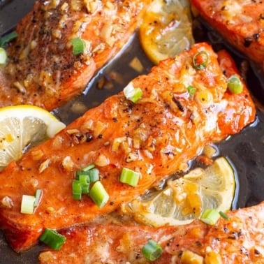 Honey garlic glazed salmon with green onion and lemon slices in cast iron skillet.