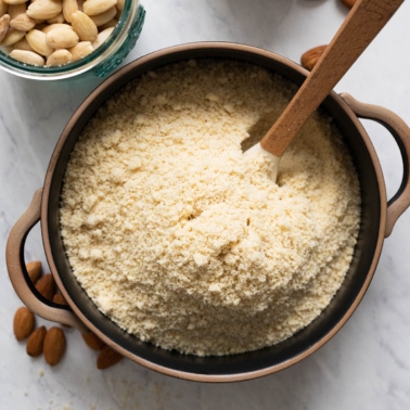 Homemade almond flour in black bowl with a spoon with blanched almonds around it.
