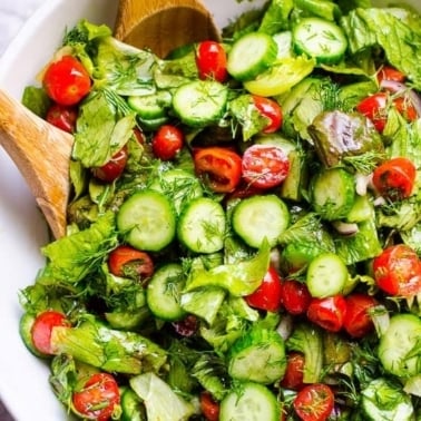 Lettuce salad with tomatoes, cucumbers and dill in white bowl with wooden spoon.
