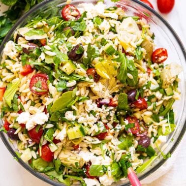 Mediterranean orzo pasta salad in a glass bowl with wooden spoon.