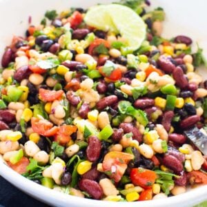 Mexican bean salad with corn, tomato, cilantro and garnished with lime in white bowl.