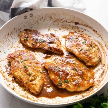 Four fried chicken breasts with sauce and garnished with parsley in a skillet.