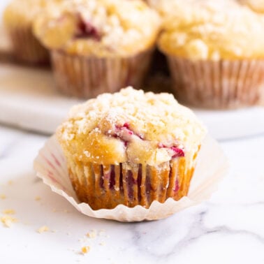 Raspberry muffin in a wrapper on a countertop and more raspberry muffins behind it.