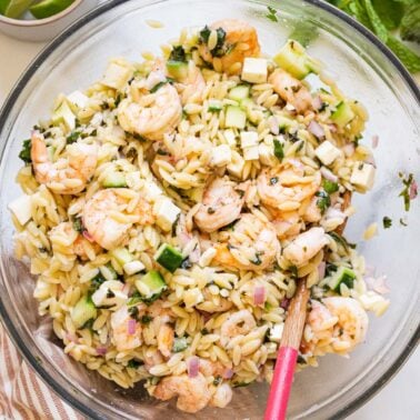 Shrimp orzo salad with cucumbers in a bowl with wooden spoon. Lime, mint and napkin on a counter.