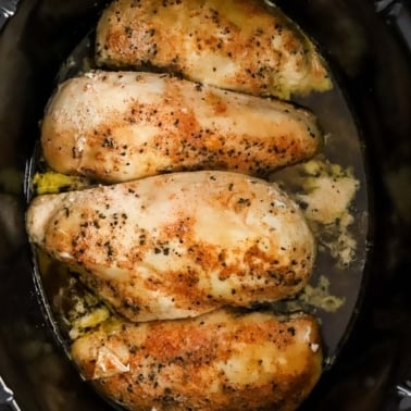Four chicken breasts in black slow cooker with seasonings in broth.