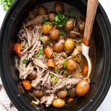 Pot roast with potatoes and carrots in slow cooker with a spoon.