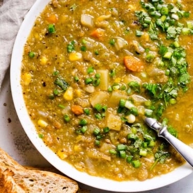Slow cooker lentil soup garnished with green onion served in a bowl with spoon. Toasted bread and linen napkin on a counter.