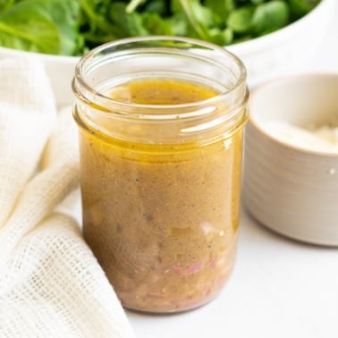Spinach salad dressing in a glass jar with spinach salad and parmesan cheese in bowls on a counter.