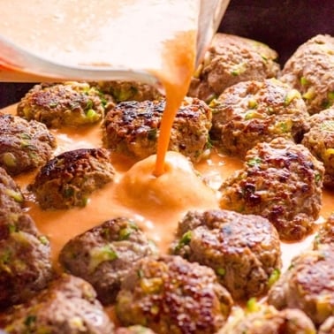 Person pouring red curry sauce over pan fried Thai meatballs in a skillet.