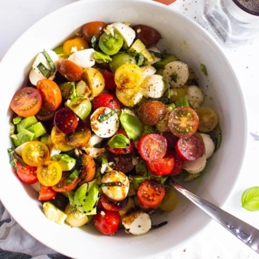 Tomato mozzarella avocado salad in a white bowl with serving spoon.