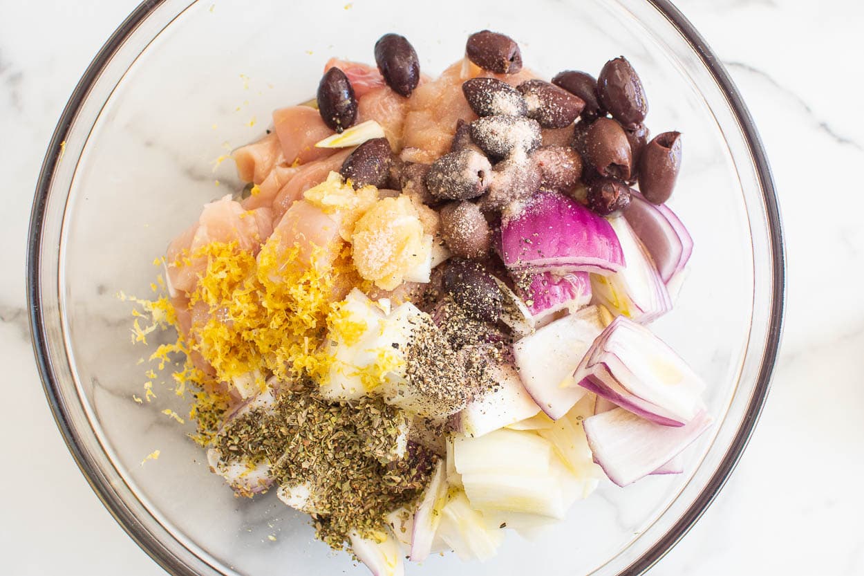 Chicken in bowl with seasonings, onion and olives.