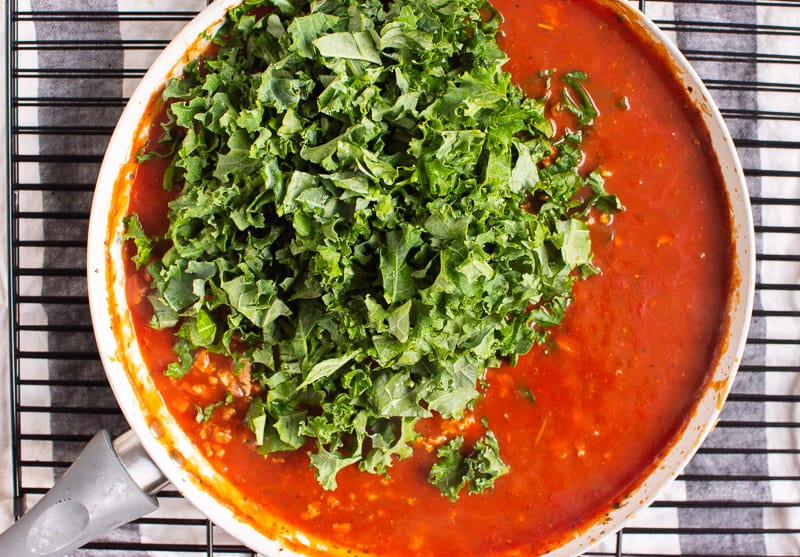 Adding in kale to tomato sauce in a skillet.