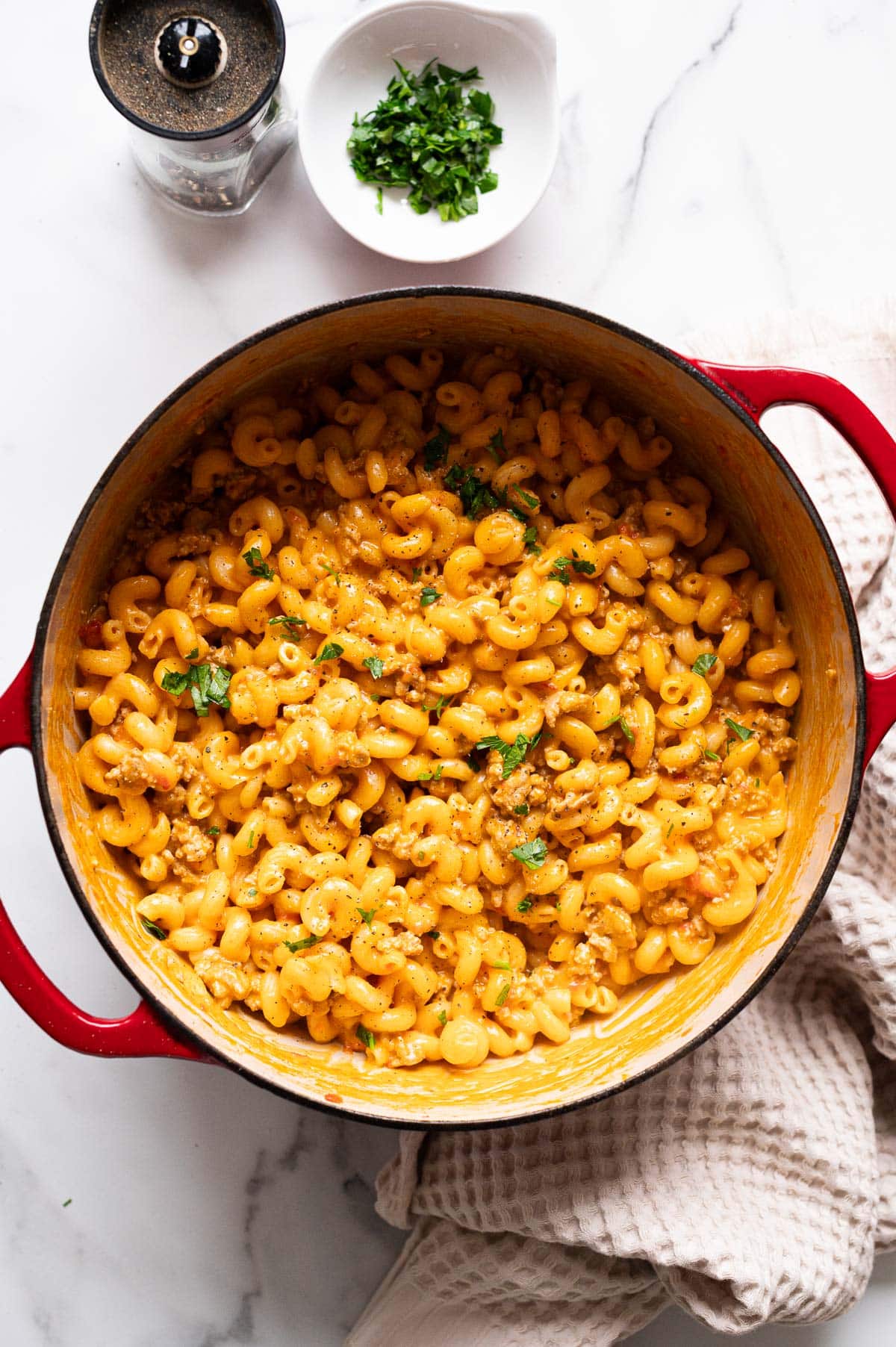 Healthy Hamburger Helper garnished with parsley in red pot. Bowl with herbs, pepper grinder and linen towel on a countertop.