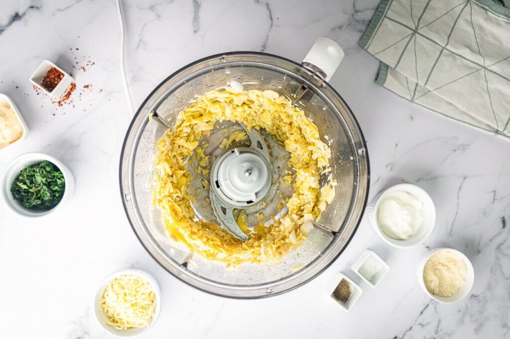 Chopped artichoke hearts, onion and garlic in a food processor. Other ingredients in small bowls around it.
