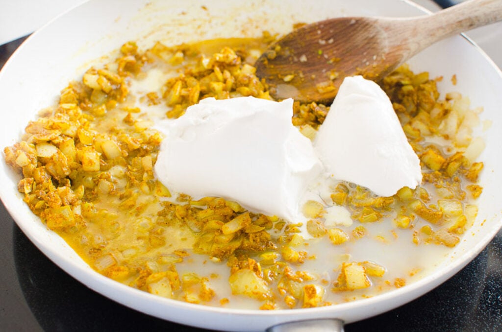 Coconut milk being stirred into curry in white skillet.