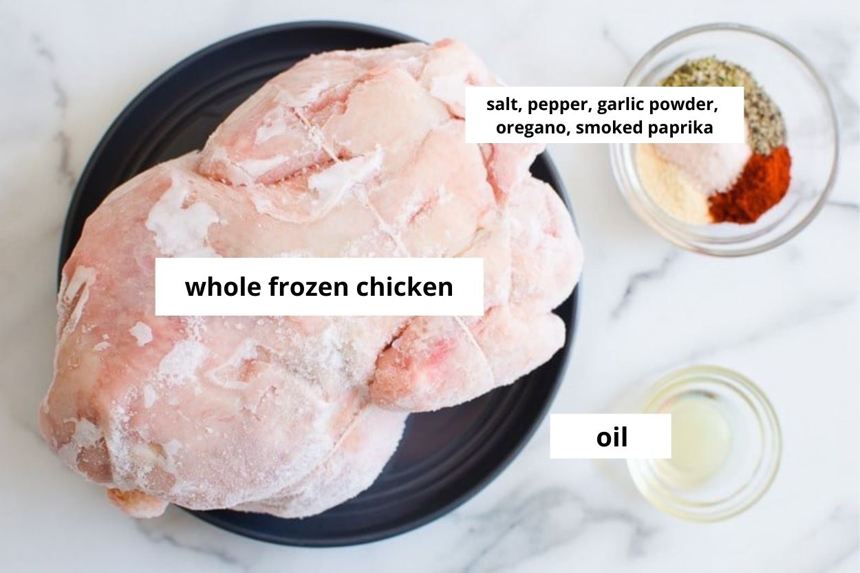 Whole frozen chicken on a plate, garlic powder, smoked paprika, dried oregano, salt and pepper and oil in bowls.