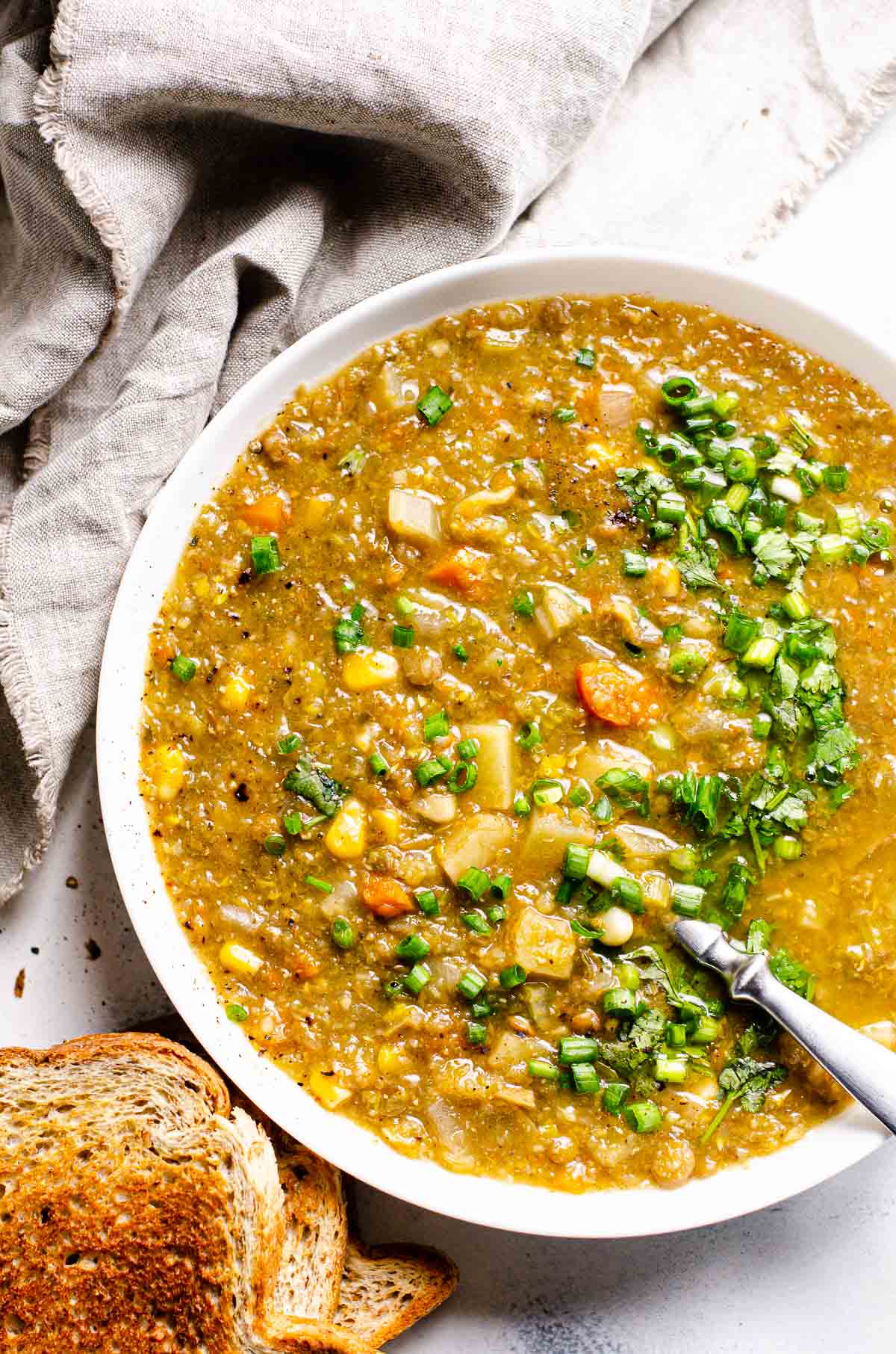 Slow cooker lentil soup garnished with green onion served in a bowl with spoon. Toasted bread and linen napkin on a counter.