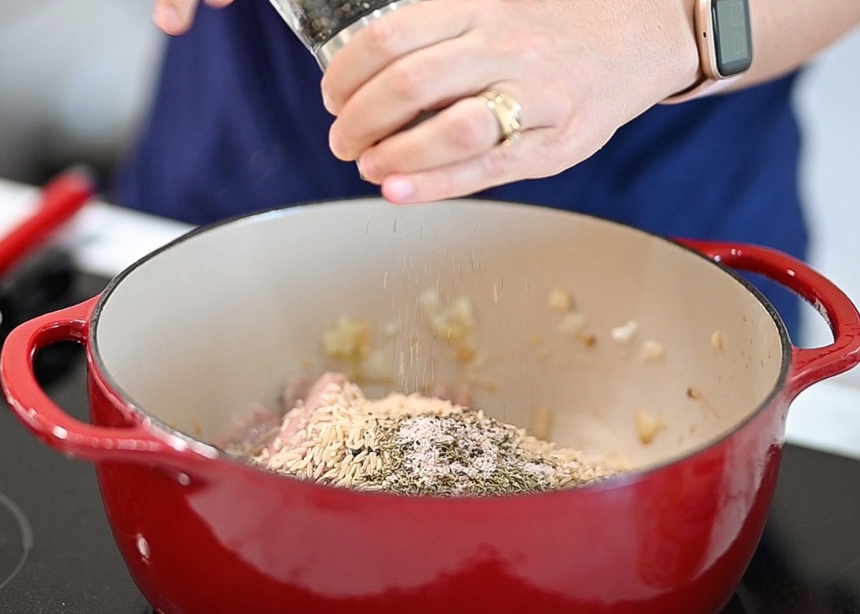 Person adding seasoning on top of meat, rice, veggies in red pot.